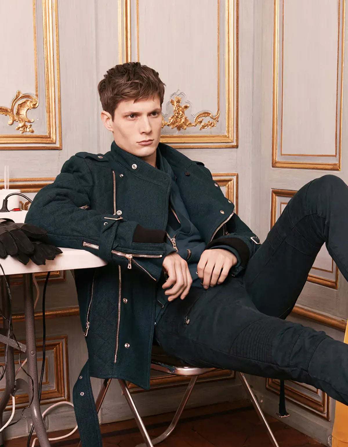 Balmain clothing for men. Man in a dark coat sitting on a chair in an elegant room with gold accents.