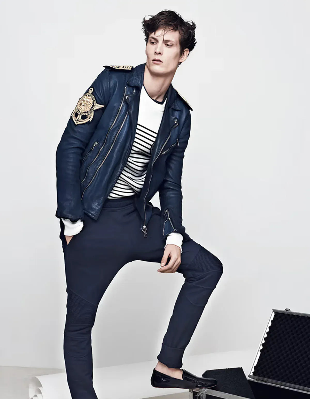 Balmain clothing for men. Man wearing a stylish outfit with a blue jacket and pants, sitting on a white surface.