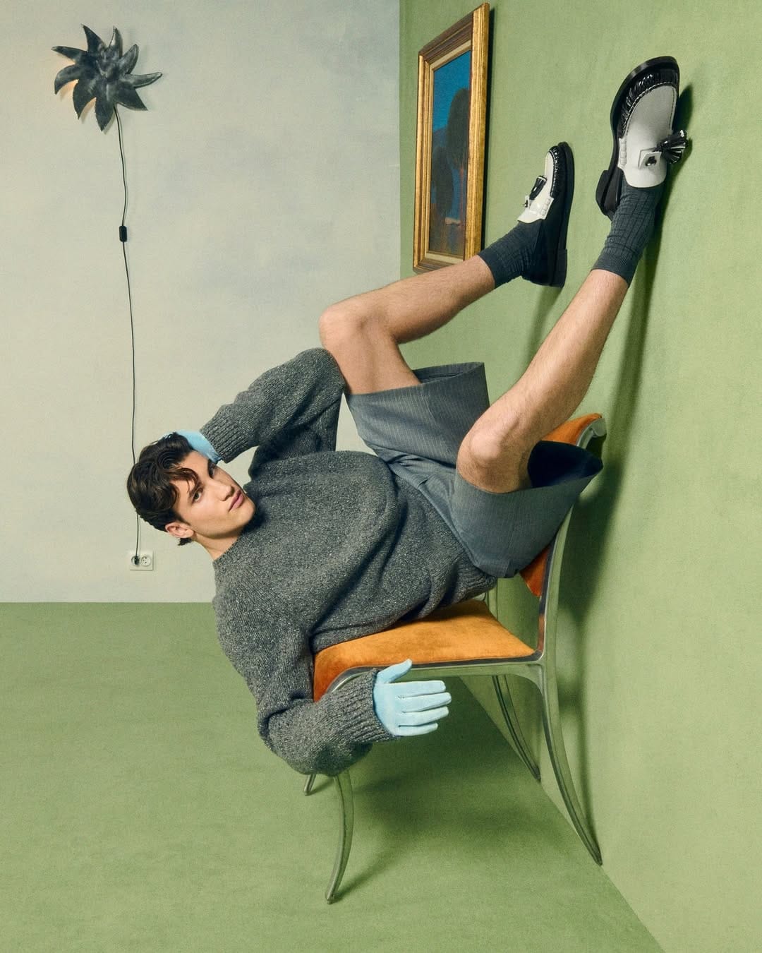 Christian Louboutin SS26. Person lying on a chair with one leg against a wall in a room with green walls and a plant.