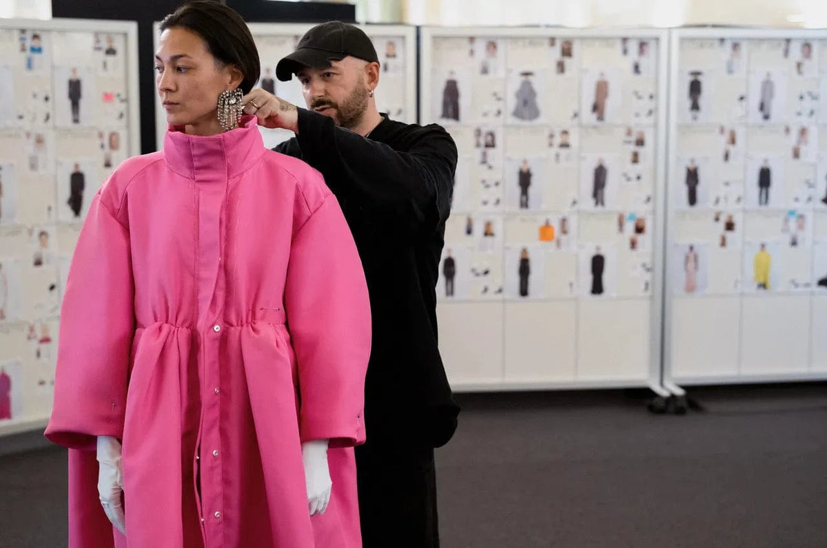 Demna Balenciaga making adjustments to the model wearing a bright pink coat, against a backdrop of fashion sketches.