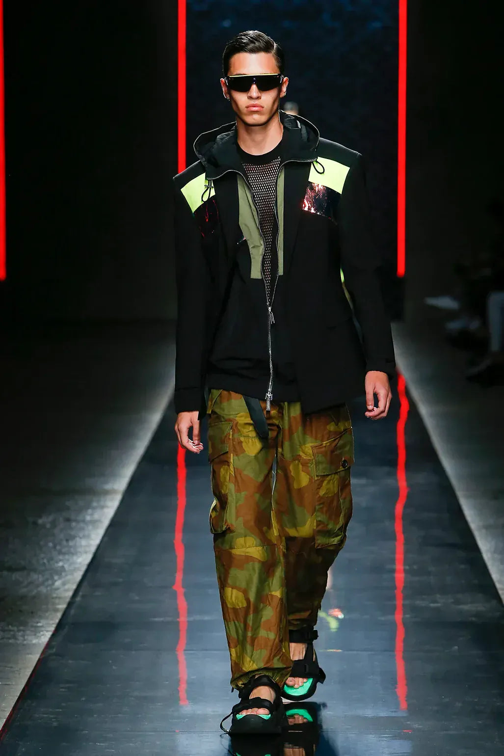 DSquared2 clothing for men. Model walking on a runway wearing a black jacket with green accents and camouflage pants.