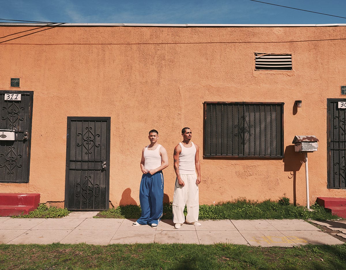 Willy Chavarria Adidas Clothing. Two men standing in front of a building with a blue sky.