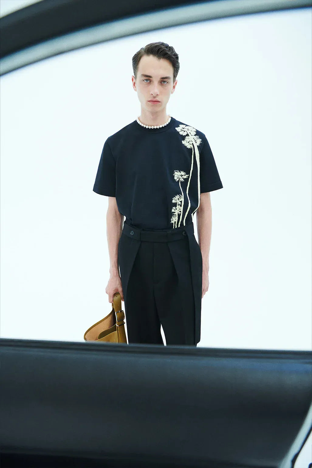Jil Sander+ Clothing For Men. Man wearing a black t-shirt with white floral designs and black pants, holding a brown bag.