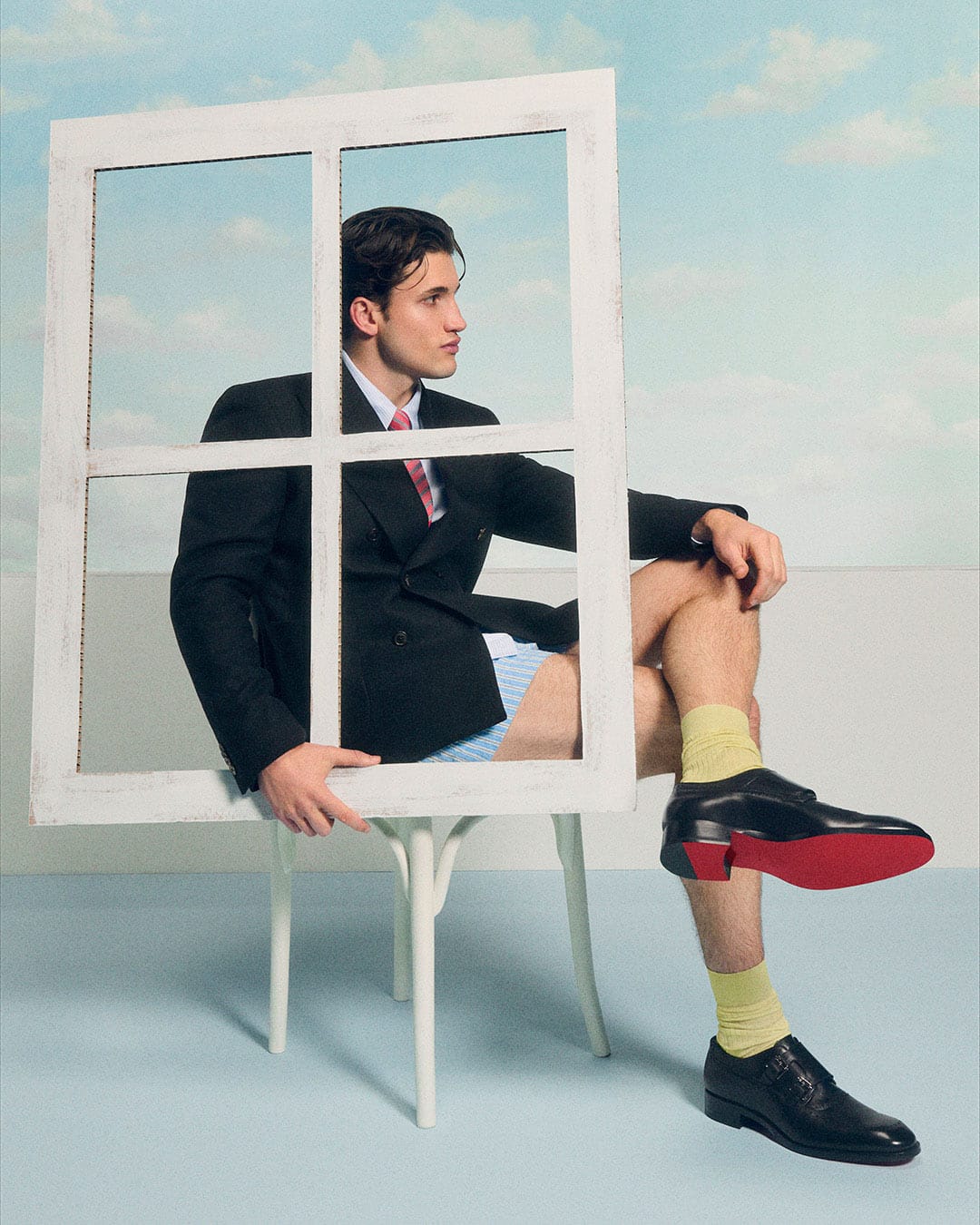 Christian Louboutin SS26. Person sitting on a chair with a window frame around them, wearing a suit and colorful socks.