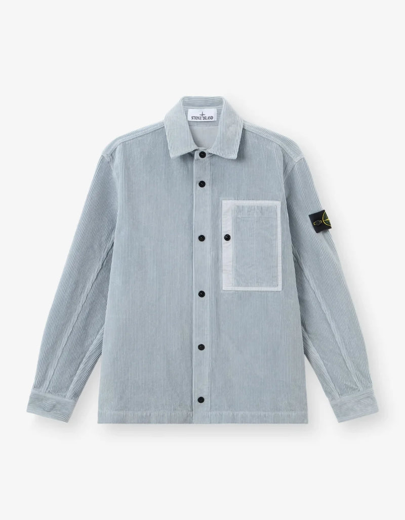 A light blue-gray Stone Island corduroy jacket, featuring the signature black and yellow compass patch on the left sleeve, set against a clean off-white background.