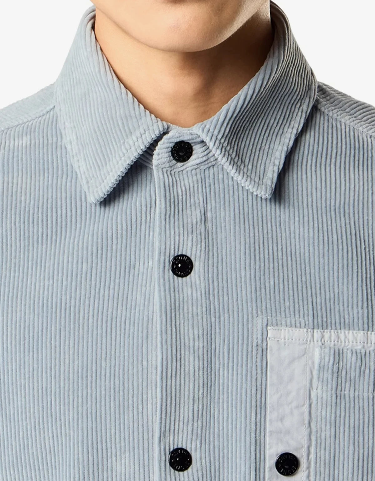 Close-up of a light blue corduroy button-up shirt with a pointed collar and black buttons, worn by a model.