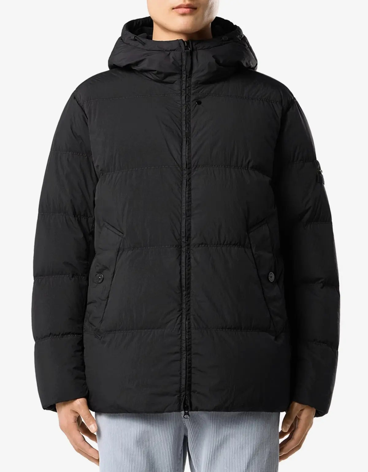 A person wearing a black hooded puffer jacket with horizontal quilting, shown from the neck down against a light gray studio background.