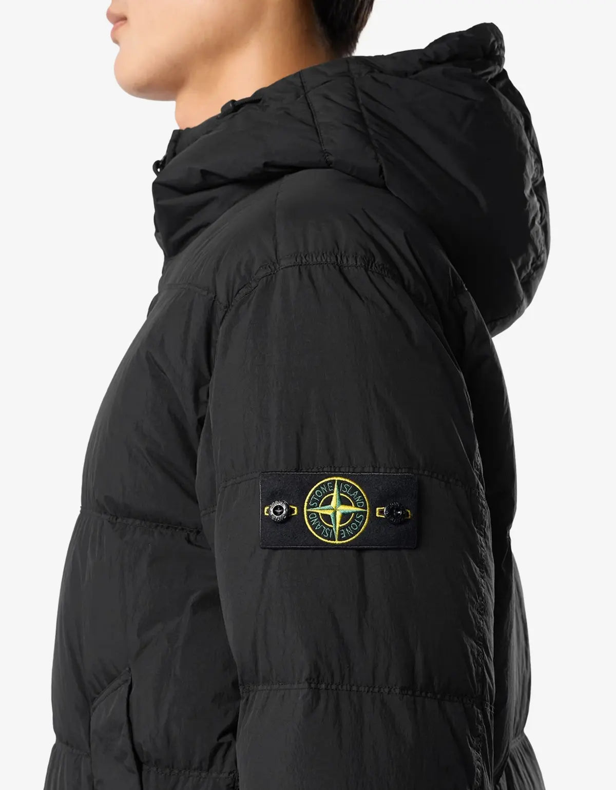 Close-up side view of a person wearing a black hooded puffer jacket with a black sleeve patch featuring the Stone Island compass logo in yellow and white.