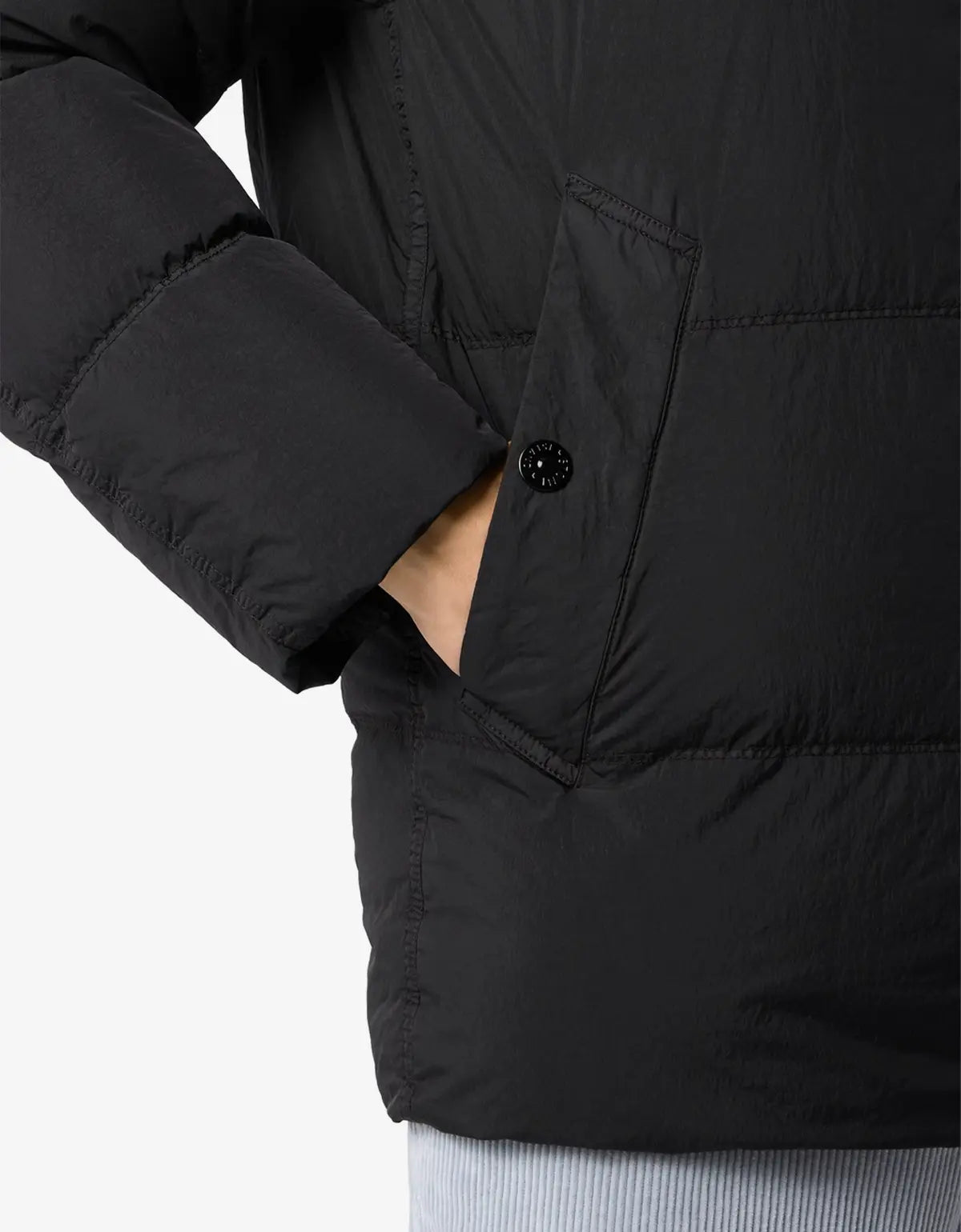 Close-up of a black puffer jacket with horizontal quilting, a side pocket with a black button, and a hand tucked into the pocket, against a light gray background.