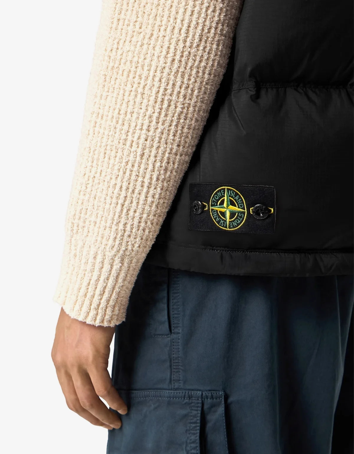 Close-up of a person wearing a black puffer vest with a Stone Island compass logo patch on the front, layered over a cream ribbed knit sweater and paired with dark blue pants.