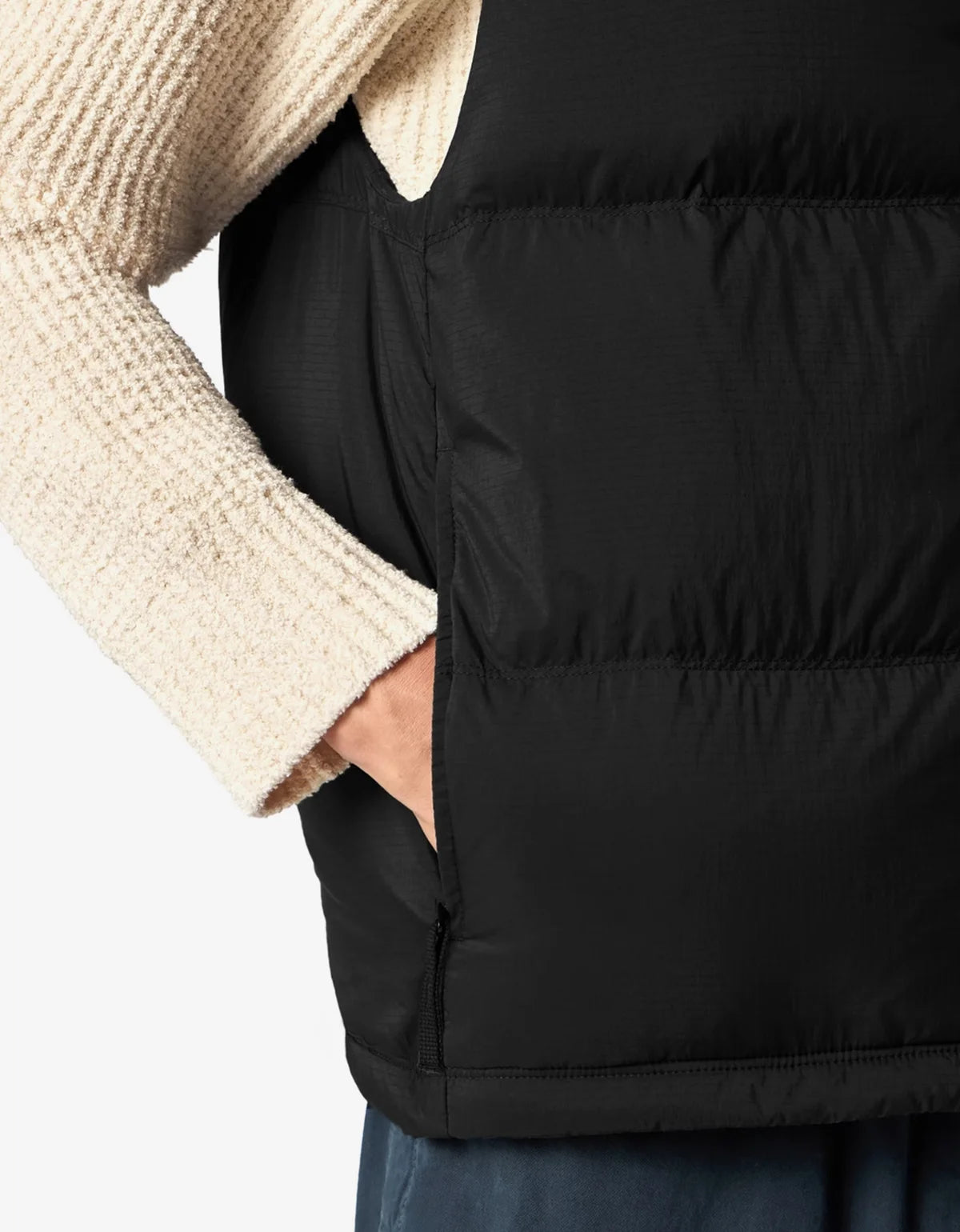 Close-up of a person wearing a black puffer vest over a cream knit sweater, with a hand in the vest's side pocket.