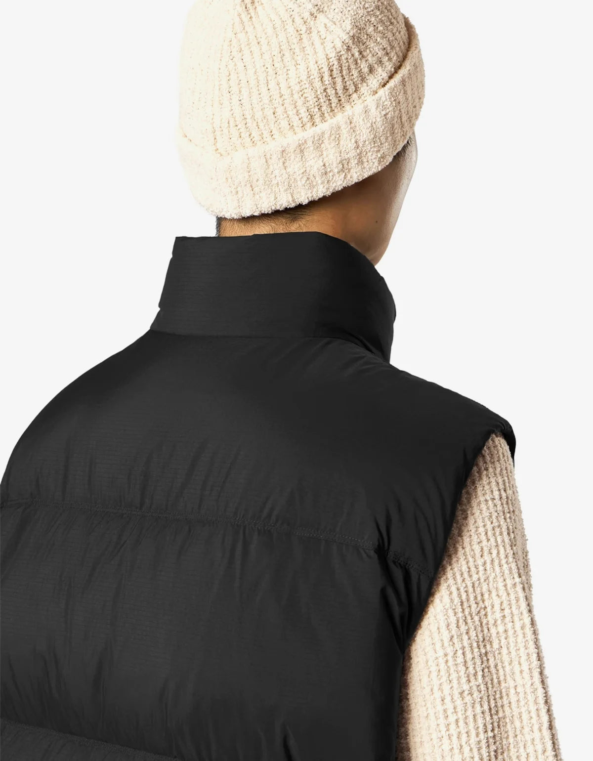 A close-up back view of a person wearing a black puffer vest over a cream knit sweater and a cream knit beanie, against a light gray studio background.