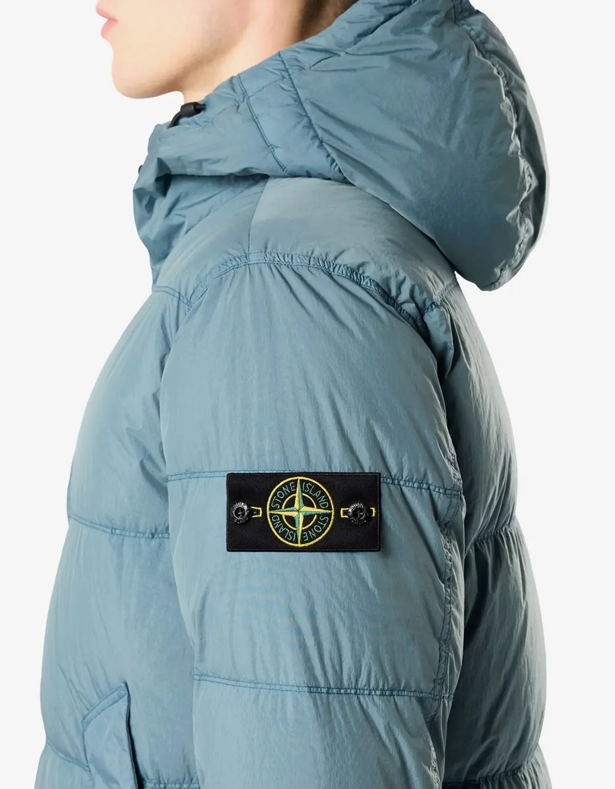 Close-up side view of a person wearing a puffer jacket in dusty blue, featuring the Stone Island compass logo patch on the upper arm.