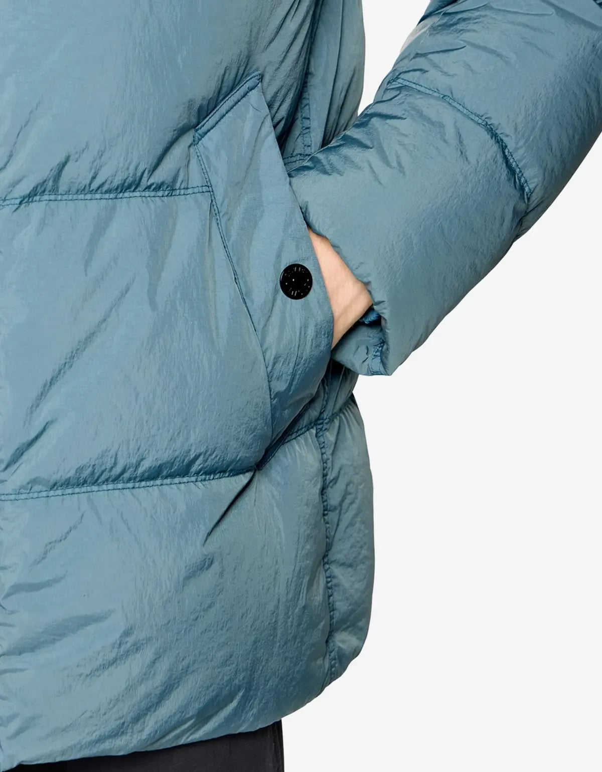 Close-up of a puffer jacket sleeve with a hand inserted into a side pocket, featuring a black button closure and a textured blue-gray fabric.
