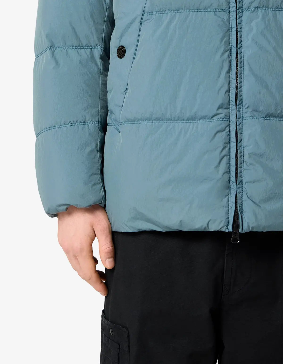 Close-up of a person wearing a muted blue-gray puffer jacket with horizontal quilting, a front zipper, and a black button, paired with black pants against a light gray background.