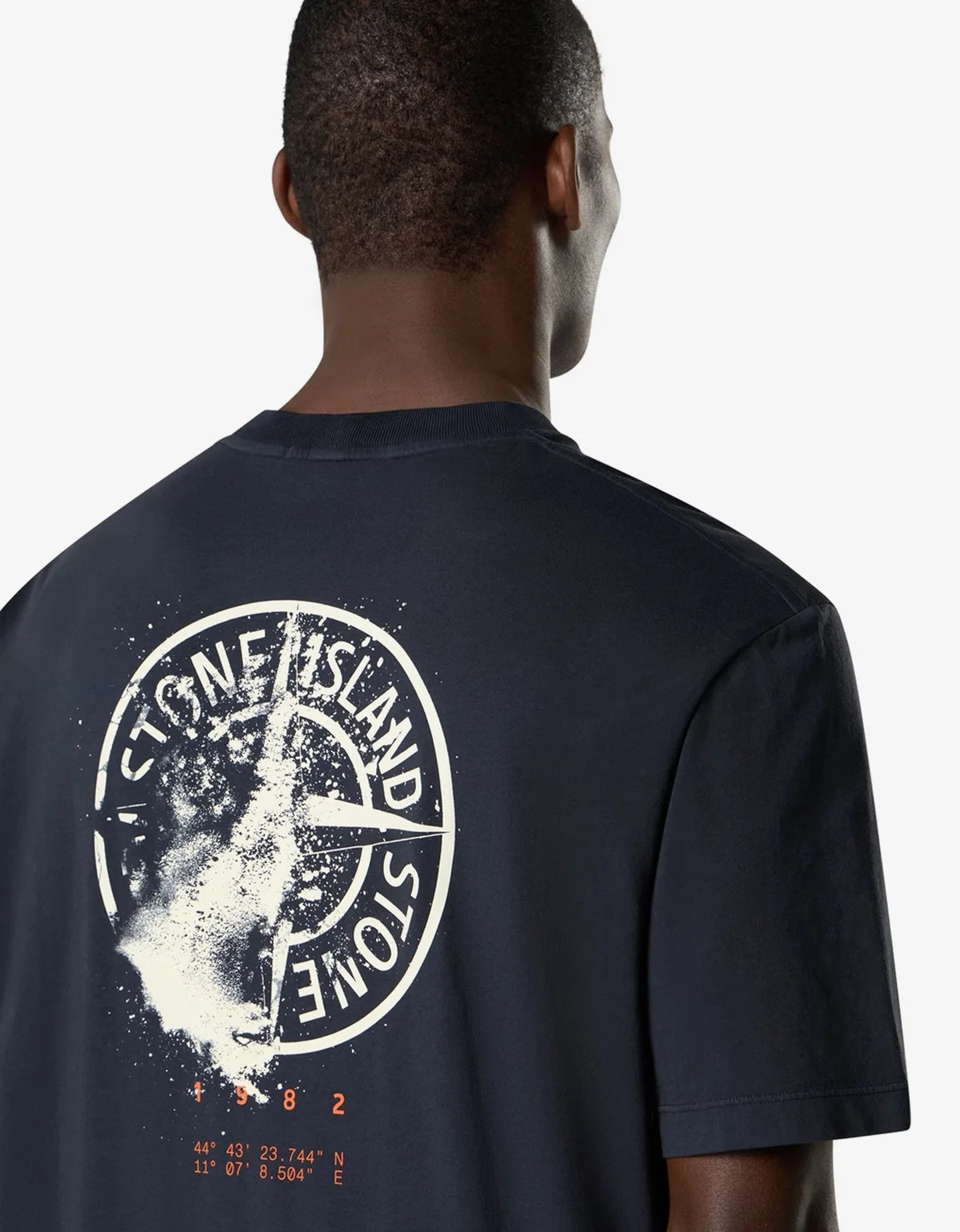 A man viewed from behind wearing a dark navy Stone Island t-shirt with a large white compass graphic and orange coordinates printed on the back.