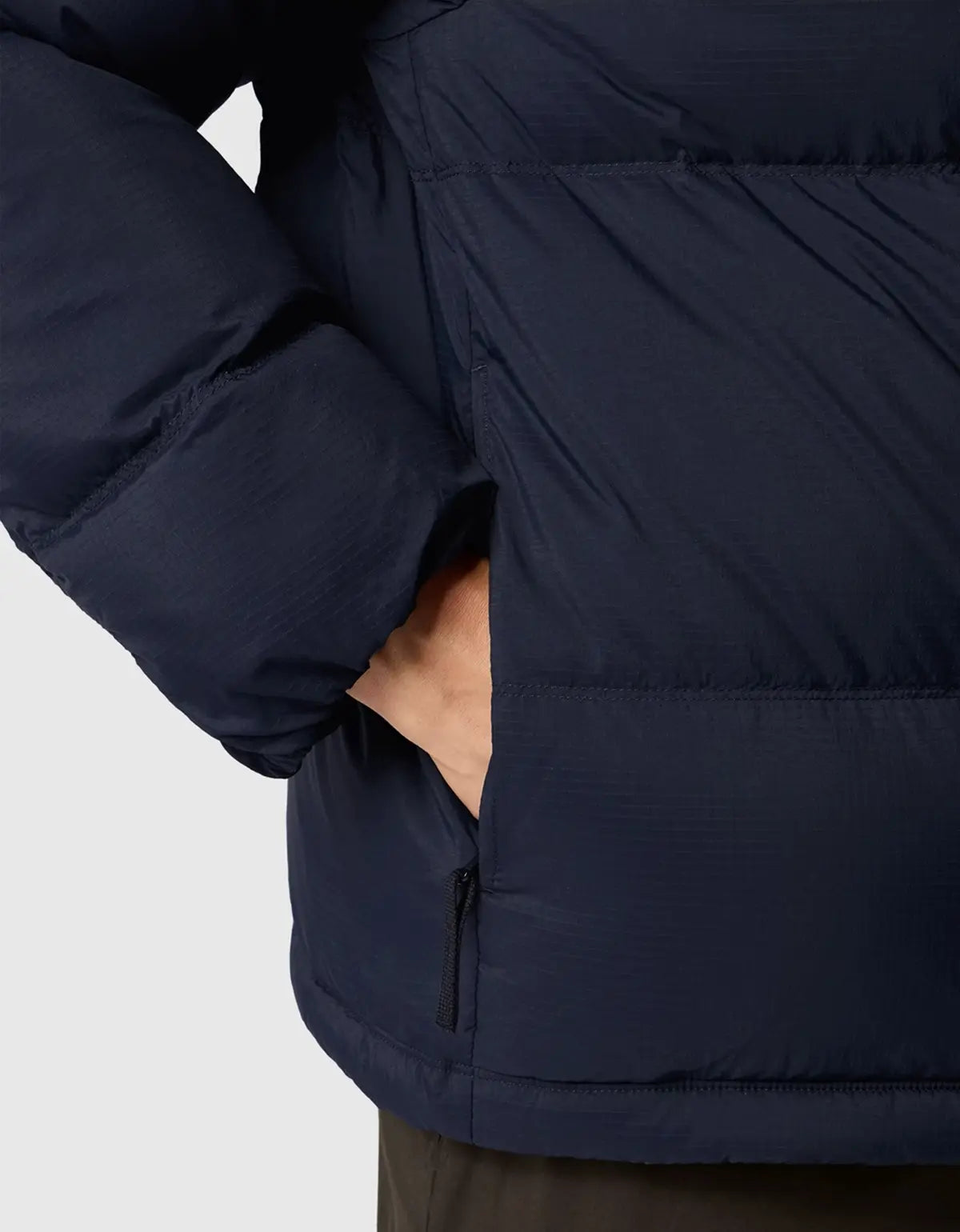 Close-up of a navy blue puffer jacket with a hand in the side pocket, showing the quilted texture and zipper detail against a light gray background.