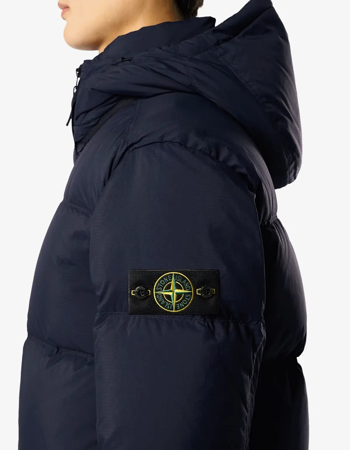 Close-up side view of a person wearing a dark navy hooded puffer jacket. The sleeve shows a black Stone Island compass patch with yellow-green detailing.