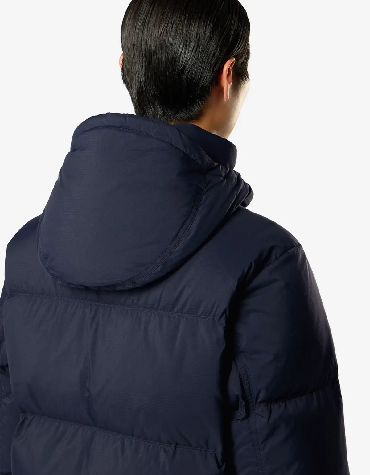 A man viewed from behind wearing a dark navy blue hooded puffer jacket against a light gray studio background.