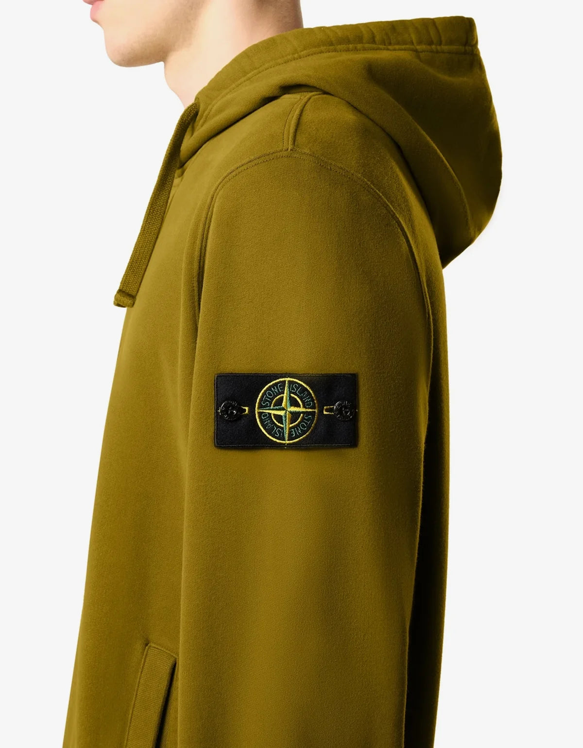 Close-up side view of a person wearing an olive green hooded sweatshirt with a black Stone Island compass patch on the upper sleeve, against a light gray background.