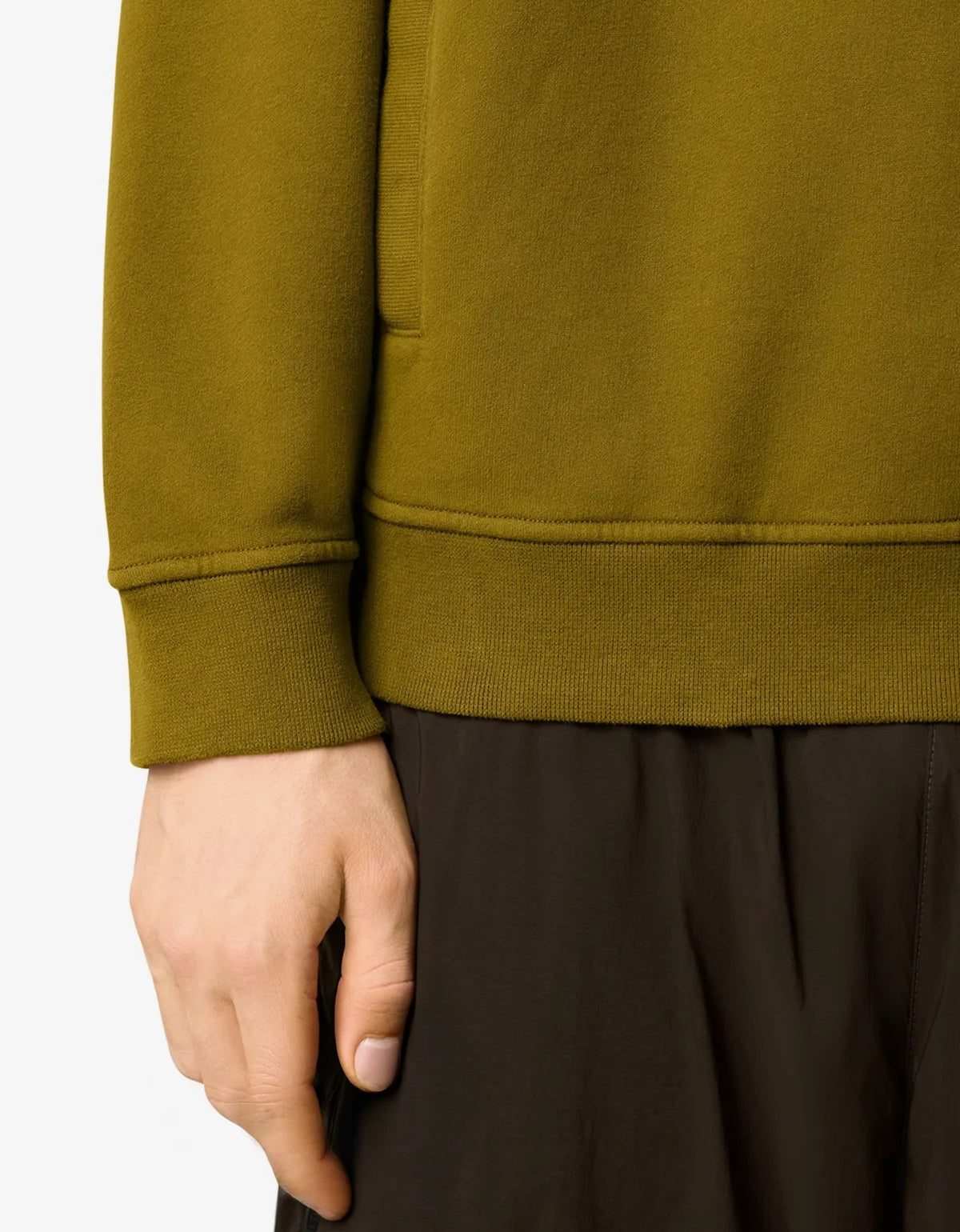 Close-up of a person wearing an olive green ribbed sweatshirt and dark brown pants, showing the sleeve cuff, waistband, and hand against a light gray background.