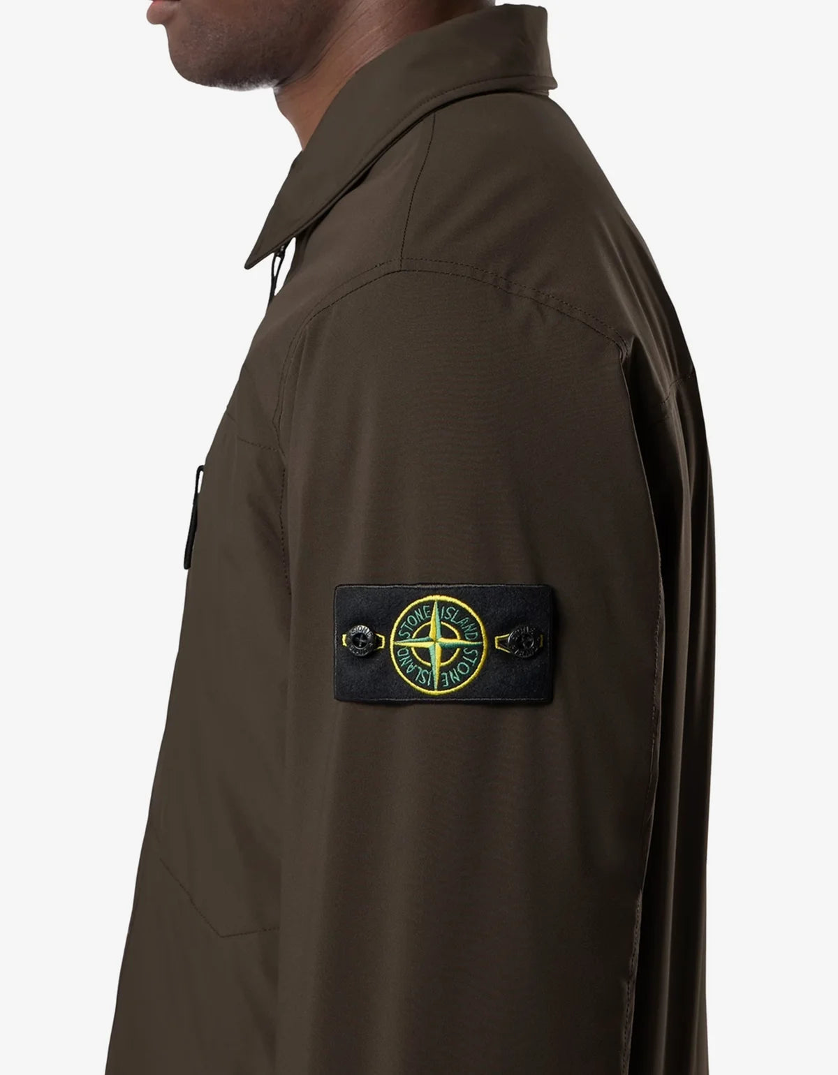 Close-up side view of a person wearing a dark olive brown Stone Island overshirt, featuring the signature compass badge on the left sleeve.