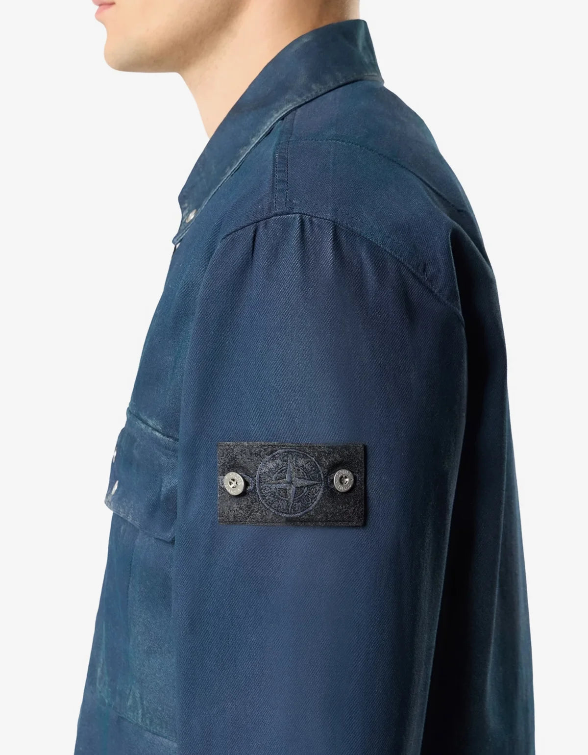 Close-up side view of a person wearing a blue Stone Island button-up shirt, showing the brand's signature compass patch on the sleeve.