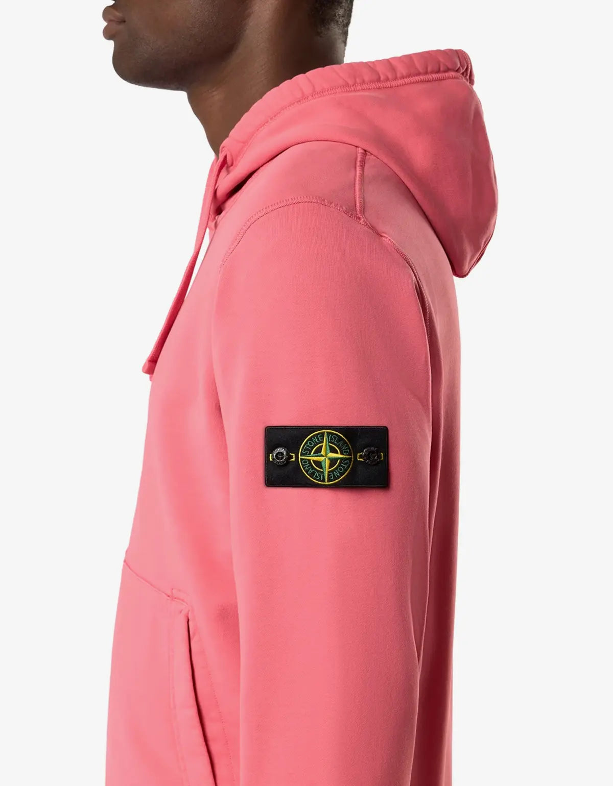 A close-up side profile of a model wearing a pink Stone Island hoodie, showing the brand's signature compass patch on the sleeve against a light gray background.
