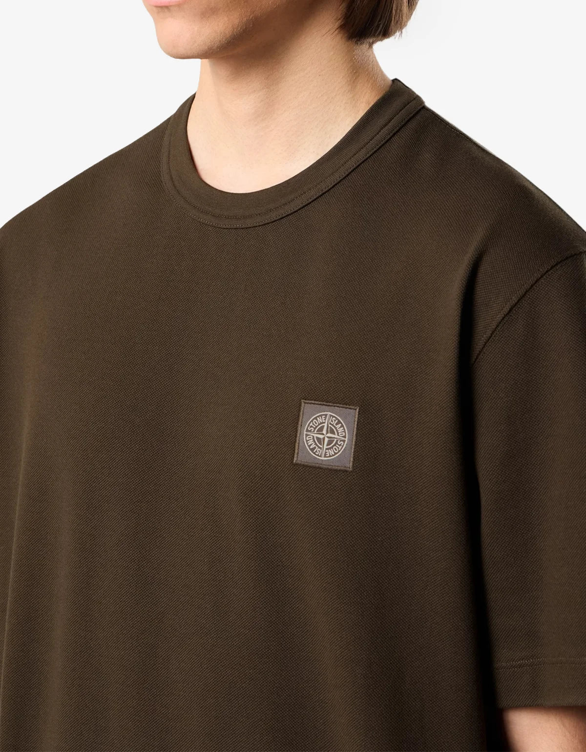 A close-up of a person wearing a dark grey crew-neck t-shirt with a small square Stone Island compass logo patch on the left chest, set against a clean white background.