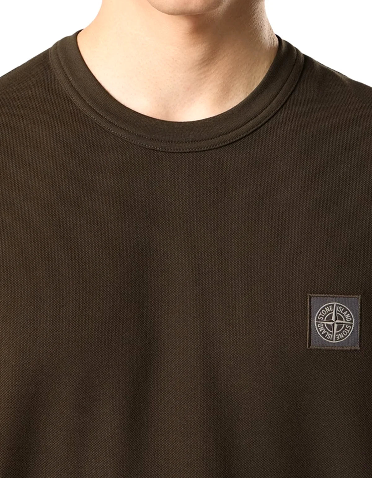 Close-up of a person wearing a dark grey crew-neck t-shirt with a small gray Stone Island logo patch on the left chest, against a plain white background.