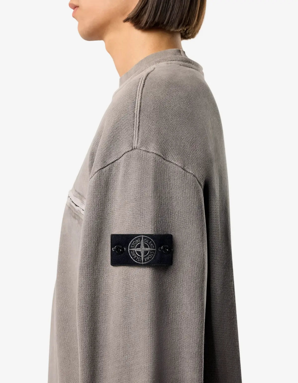 Close-up side view of a person wearing a gray knit sweater with a black Stone Island compass patch on the sleeve, against a light gray background.