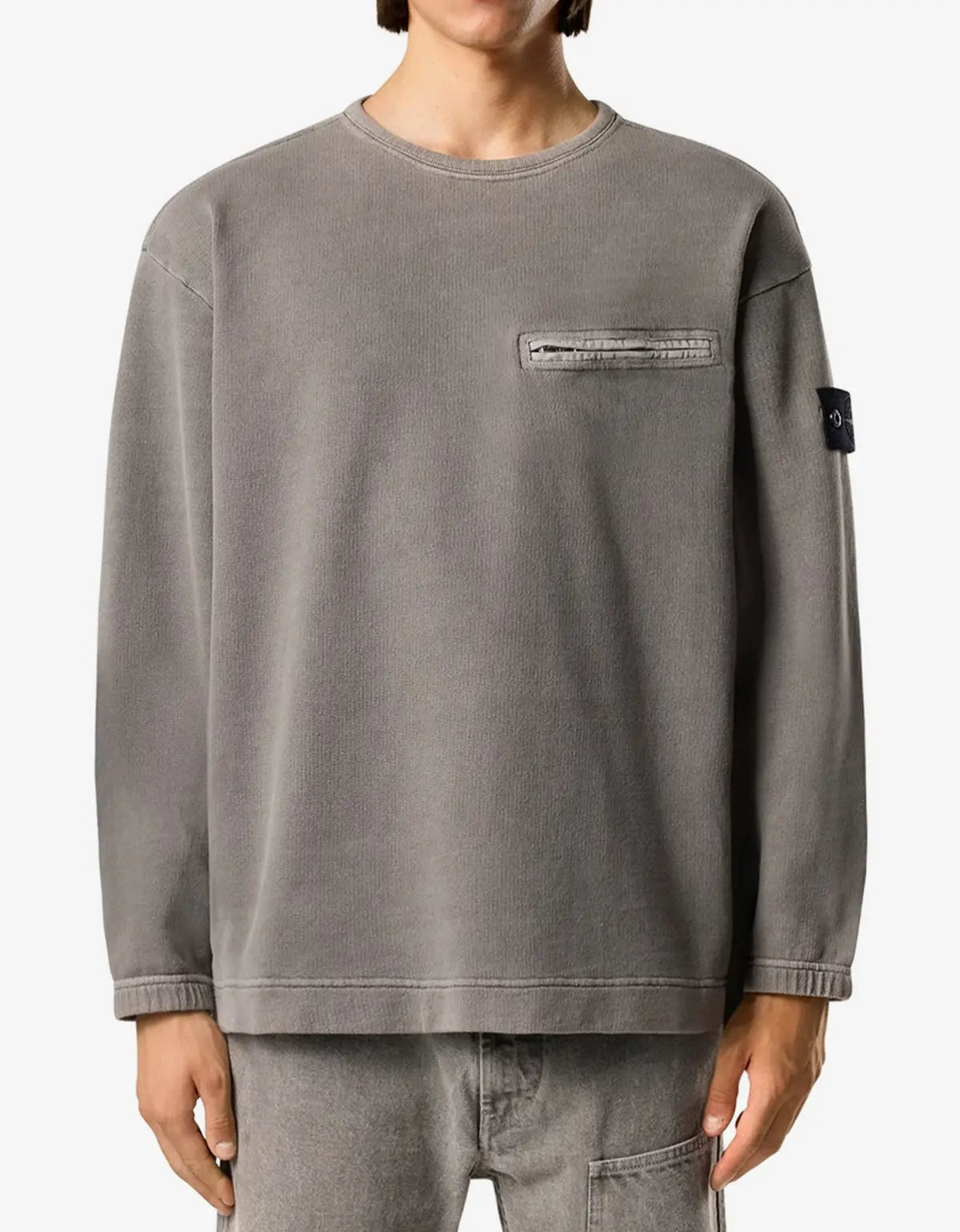 A person wearing a gray crewneck sweatshirt with a zippered chest pocket and a small black brand patch on the sleeve, paired with gray jeans against a plain light background.