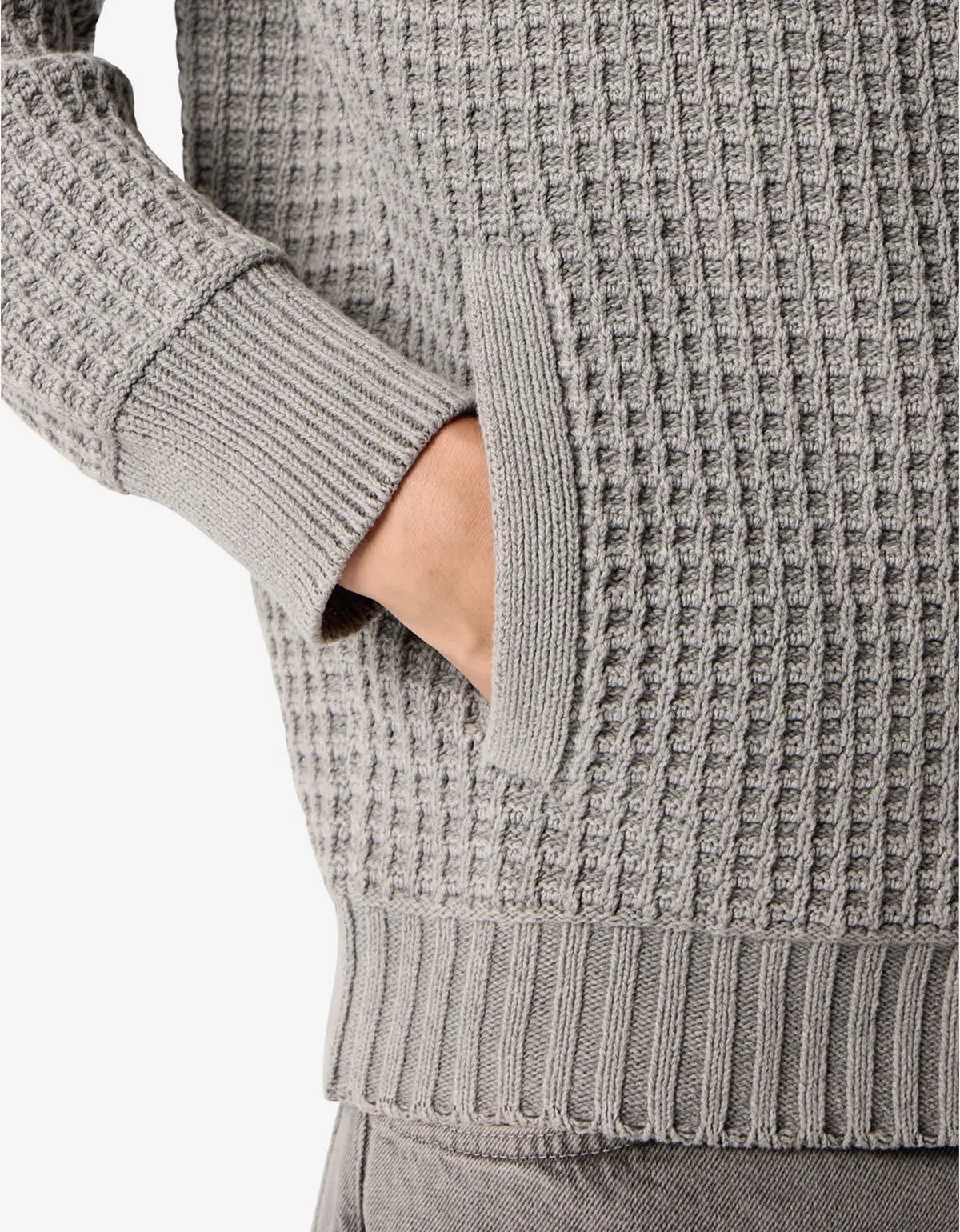 Close-up of a gray waffle-knit sweater with a ribbed cuff, pocket, and hem, shown on a model with one hand in the pocket.