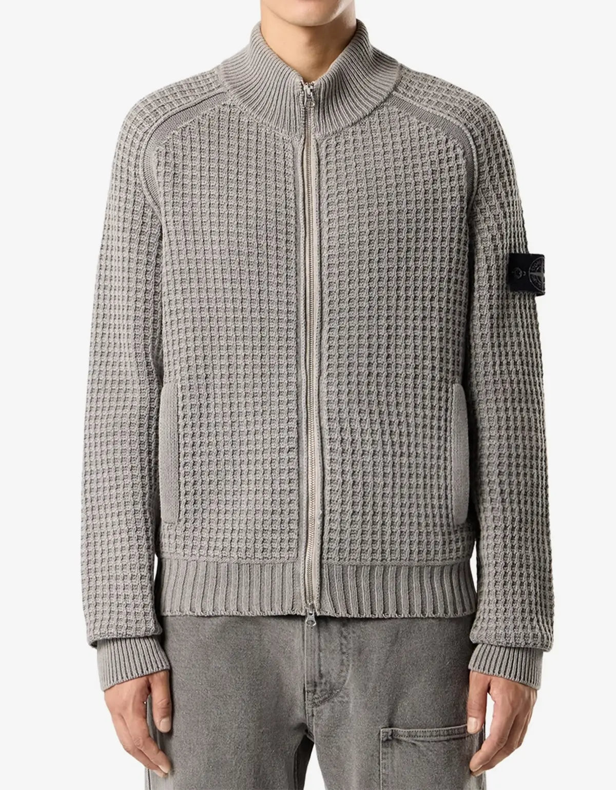 A gray waffle-knit zip-up jacket with a mock neck, ribbed cuffs and hem, featuring a black Stone Island brand patch on the left sleeve, worn by a model.
