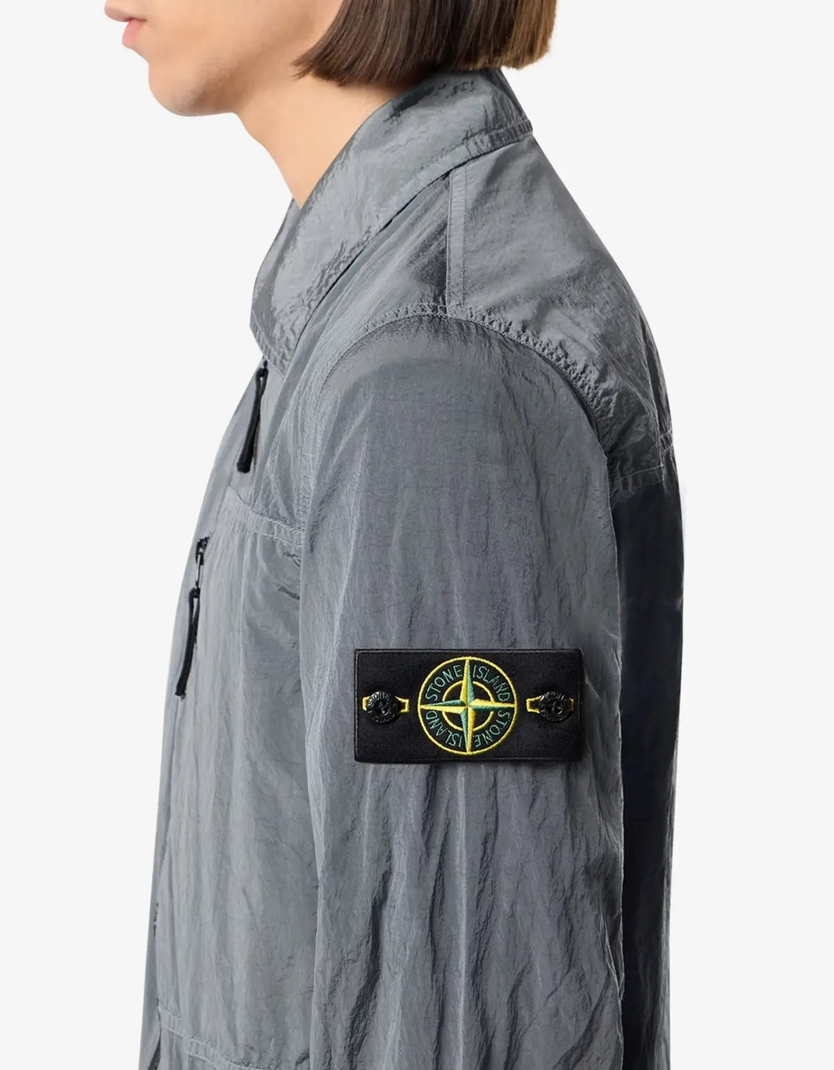 A close-up side profile of a person wearing a gray lightweight jacket with a black Stone Island compass patch on the sleeve, against a plain light gray background.