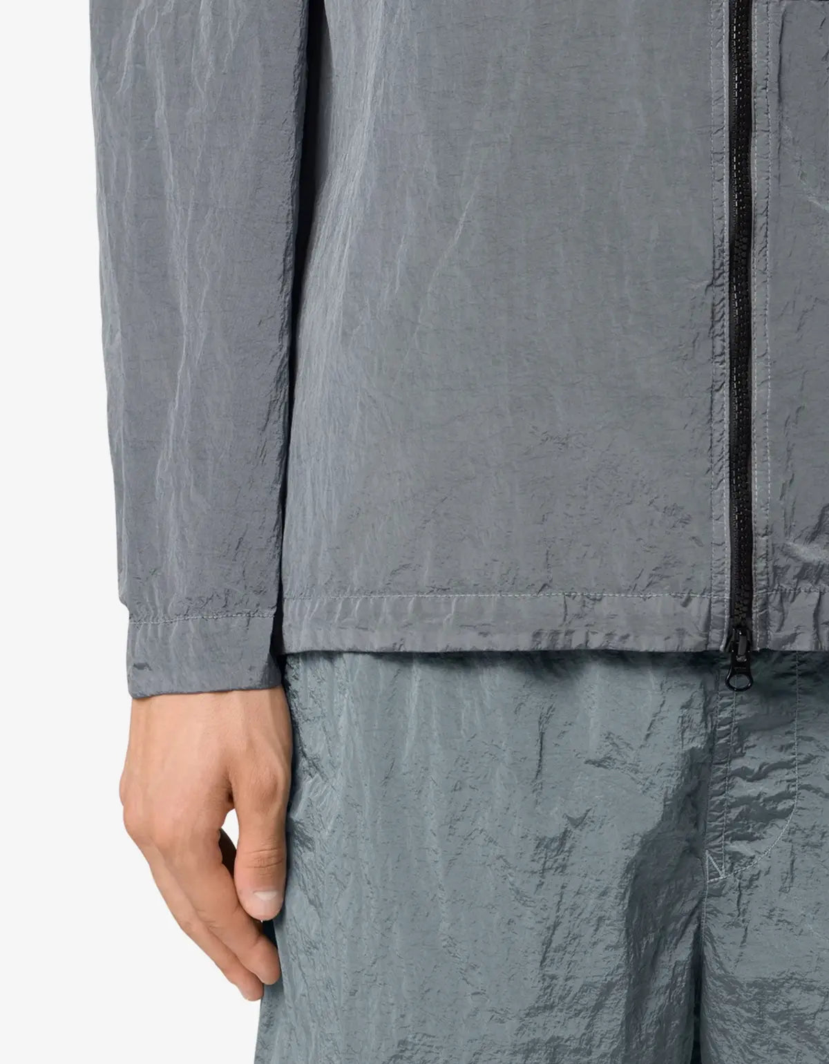 Close-up of a person wearing a gray crinkled lightweight jacket with a black front zipper and matching gray crinkled pants, cropped to show the torso and hand against a plain light background.