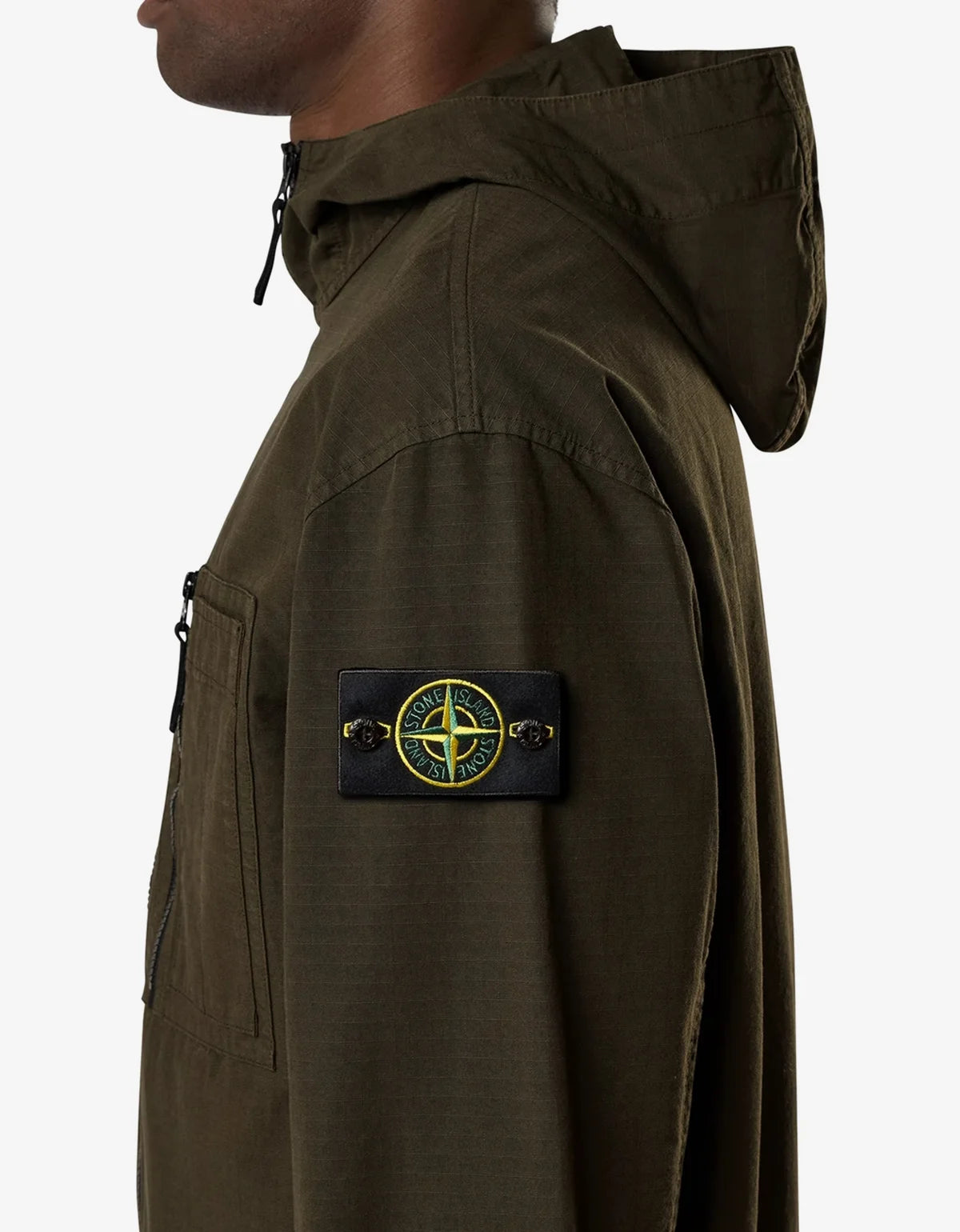 Close-up side view of a person wearing an olive green hooded jacket with a black Stone Island compass patch on the sleeve, against a light gray background.