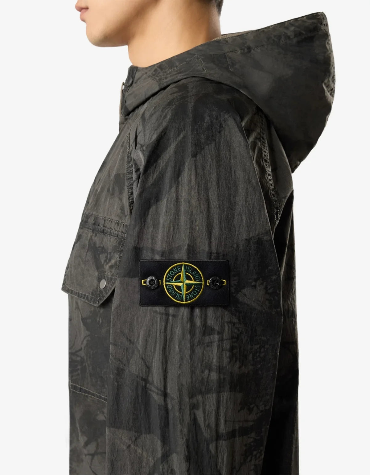 Side profile of a model wearing a Stone Island hooded jacket in a dark gray and black camouflage pattern, featuring the signature compass patch on the sleeve.