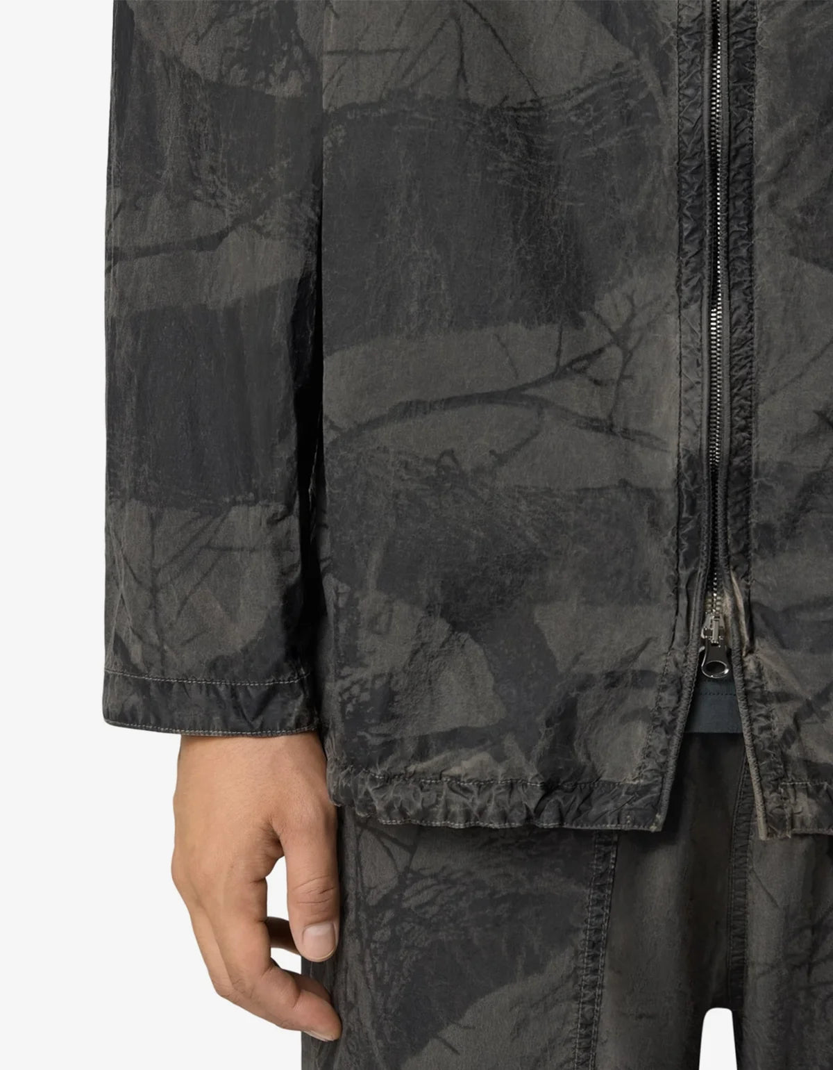 Close-up of a person wearing a dark gray camouflage jacket with a front zipper and matching dark pants against a plain light background.