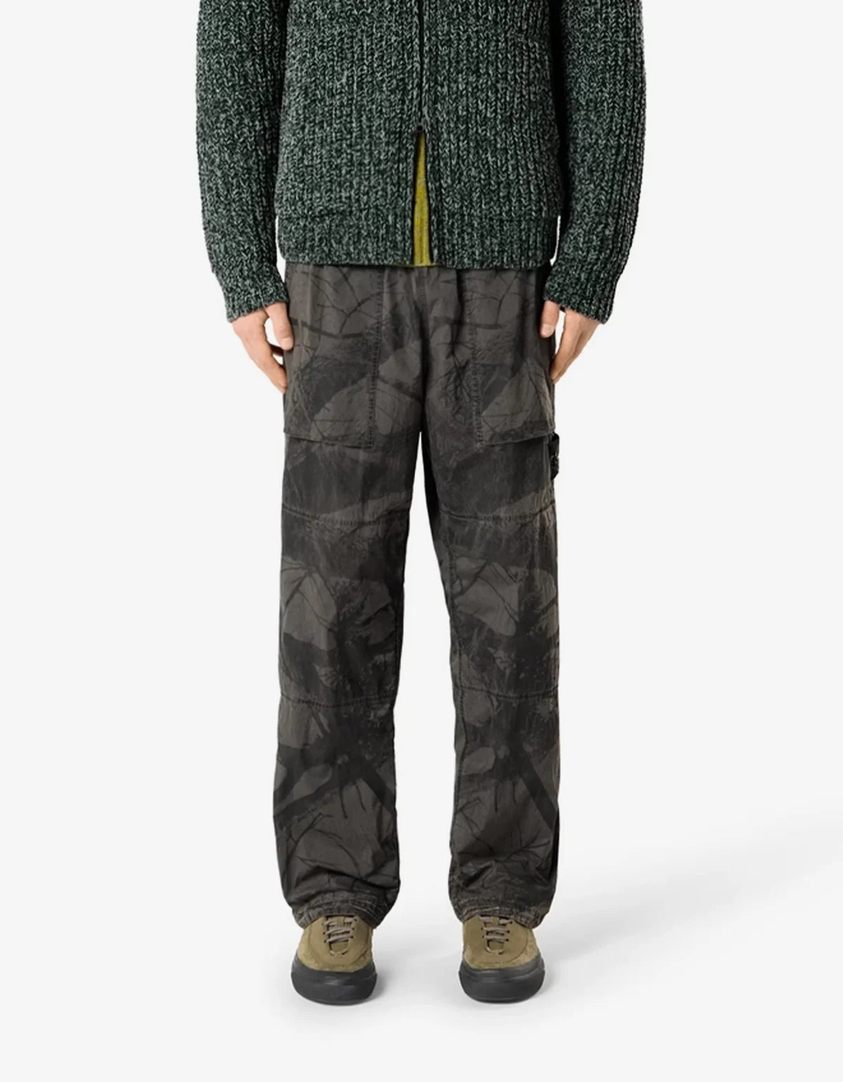 A person wearing dark camouflage cargo pants with a dark green textured knit sweater and olive shoes, shown from the chest down against a light gray studio background.