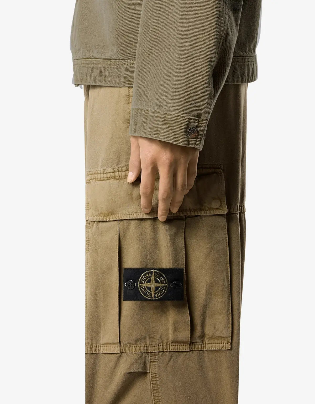 Close-up of a person wearing tan cargo pants with a large front flap pocket and a black Stone Island compass logo patch, against a light gray background.