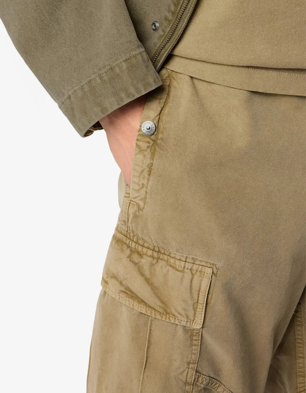 Close-up of a person wearing tan canvas utility pants with a side pocket and a silver button, paired with a matching tan zip jacket, against a plain white background.