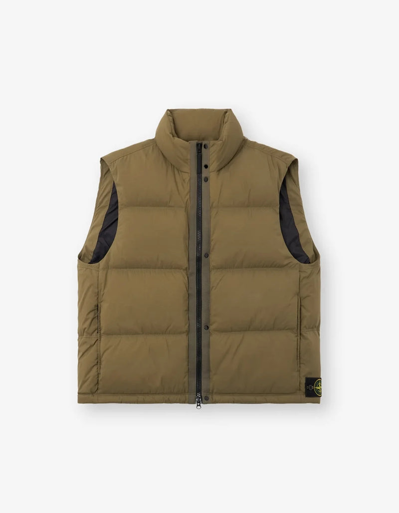 A sleeveless puffer vest in olive green with black side panels and a black front zipper, shown flat against a light gray background. A small Stone Island compass logo is visible on the bottom right hem.
