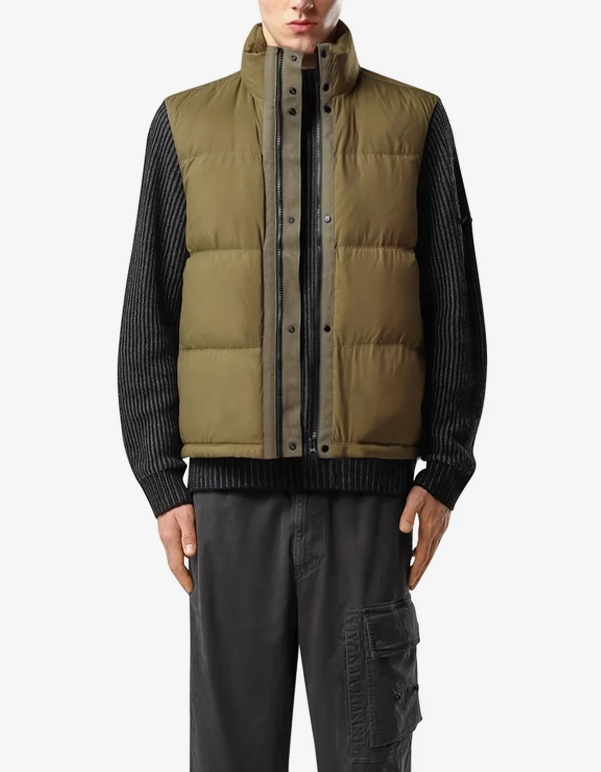 A model wearing a sleeveless olive green puffer vest over a black ribbed knit sweater and dark gray cargo pants, shot against a light gray studio background.
