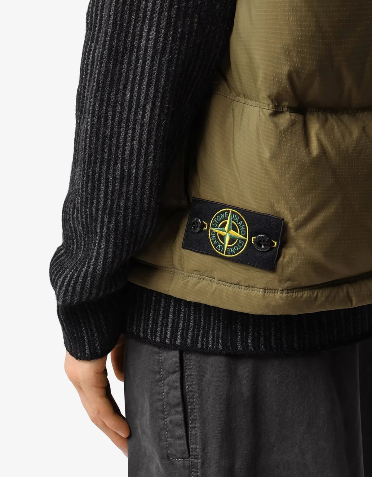 Close-up of a person wearing an olive green puffer vest over a dark gray ribbed sweater, with a Stone Island compass logo patch visible on the back.