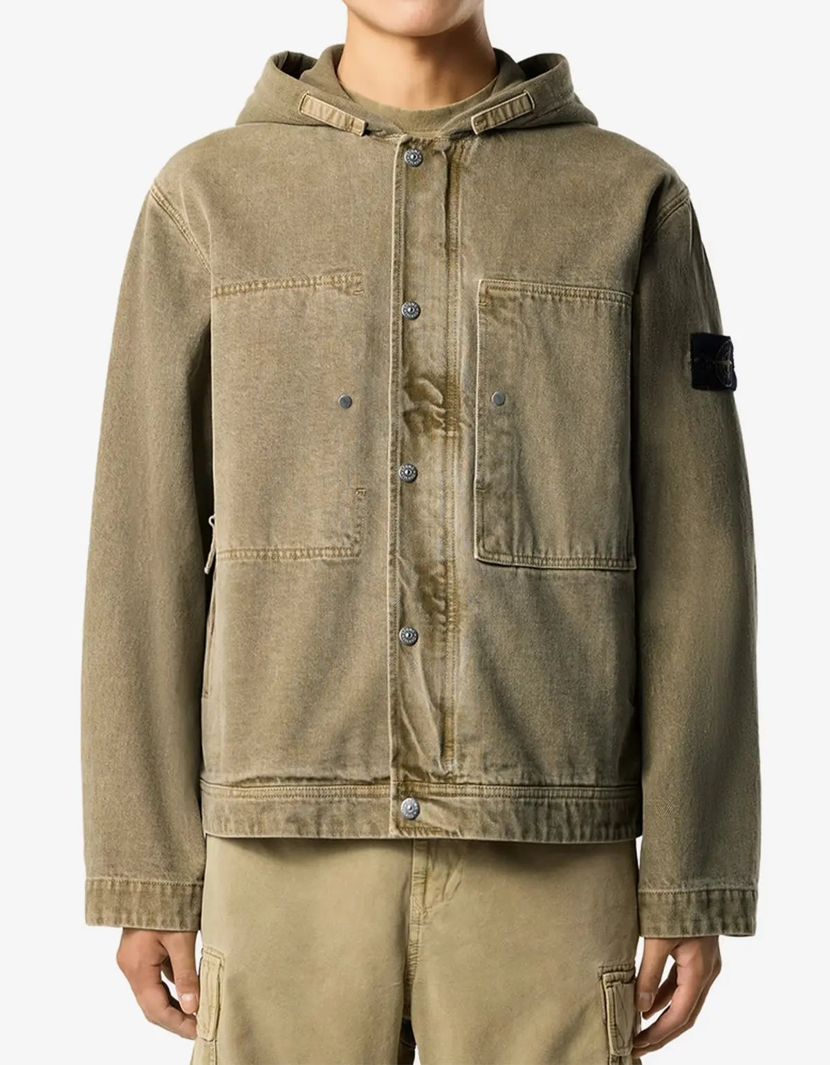 A person wearing a muted olive-brown hooded jacket with front buttons, flap chest pockets, and a black rectangular patch on the upper sleeve, paired with matching pants against a plain light background.