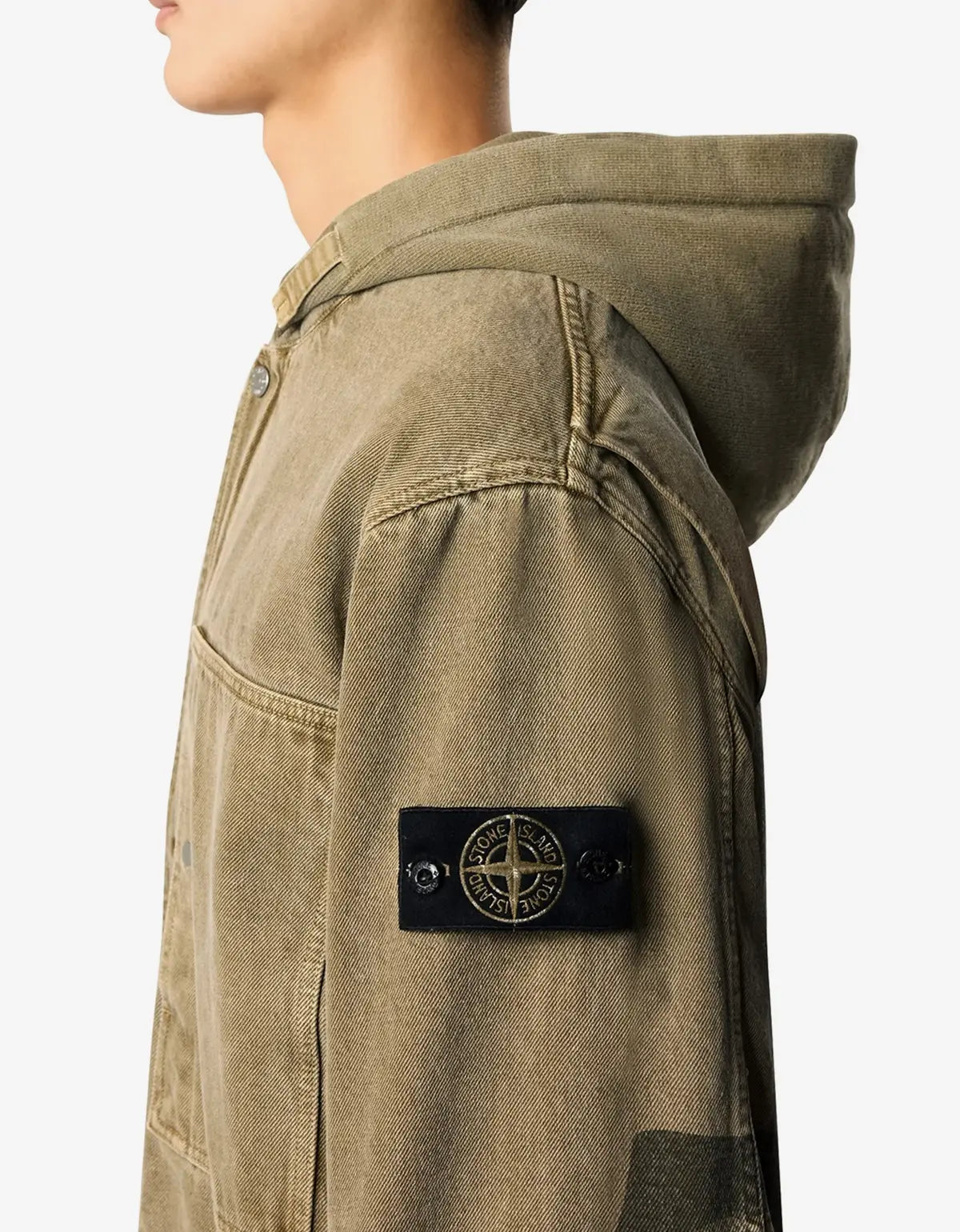 Close-up side view of a person wearing an olive green hooded jacket with a black Stone Island compass patch on the sleeve, against a light gray background.