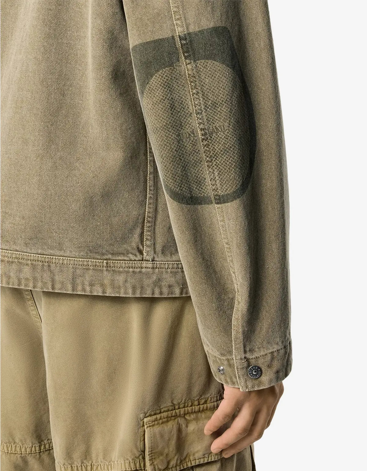 Close-up of a person wearing a washed olive-brown jacket with a dark gray patch on the upper sleeve and matching cargo pants against a light gray background.