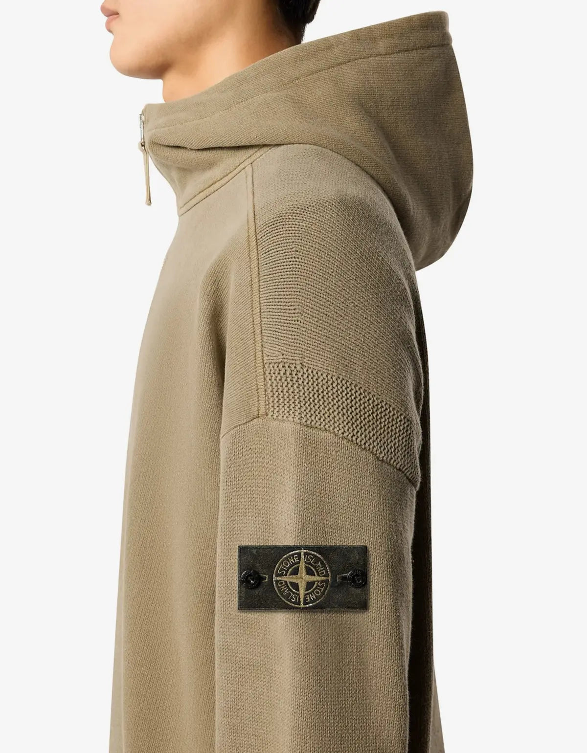 Close-up side view of a person wearing a beige hooded pullover with textured knit shoulder panels and a dark sleeve patch featuring the Stone Island compass logo.