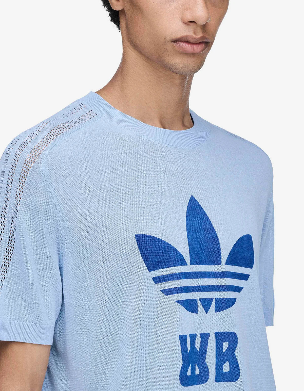 A close-up of a model wearing a light blue short-sleeve t-shirt with a dark blue Adidas Trefoil logo and 'WB' initials printed on the front, featuring mesh detailing on the shoulder.